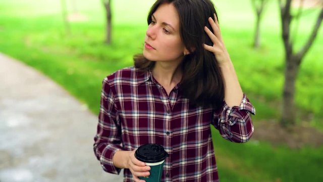 Young pretty girl standing on path in the park and drinking a cup of coffee