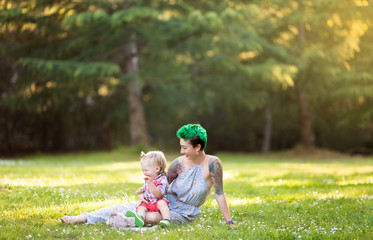 Young mother with green short hair is sitting on green grass, smiling, looking at her toddler son on her laps. Mother's day concept	