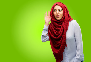 Young arab woman wearing hijab holding hand near ear trying to listen to interesting news...