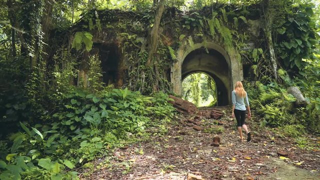 Young caucasian woman walking through gate of old Fort Karang Bolong covered with tree roots, ferns and vines in Cilacap, Indonesia