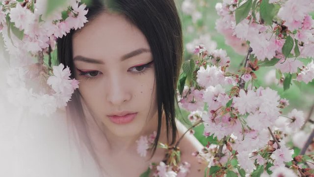 Outdoor fashion concept shoot of beautiful young asian woman surrounded by flowers on spring. Perfect model with creative vivid makeup and pink lipstick on lips and traditional japanese hairstyle