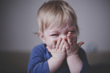 Cute little blond toddler in blue clothes bitterly cries and presses his hands to his face. Close...