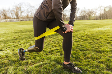 Cropped photo of disabled girl in tracksuit, exercising and doing lunges with prosthetic leg on grass using resistance band