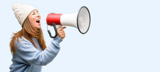 Middle age woman wearing wool winter cap communicates shouting loud holding a megaphone, expressing success and positive concept, idea for marketing or sales isolated blue background
