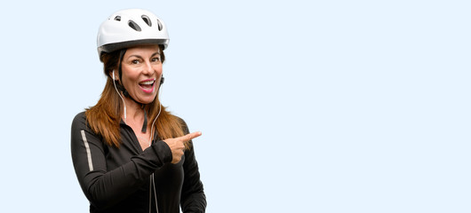 Middle age cyclist woman using earphones pointing away side with finger isolated blue background