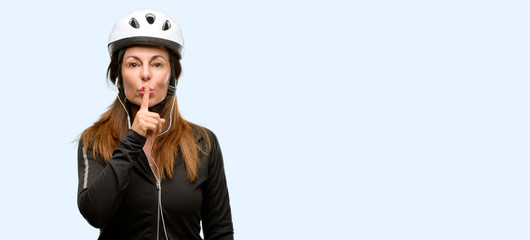 Middle age cyclist woman using earphones with index finger on lips, ask to be quiet. Silence and secret concept isolated blue background