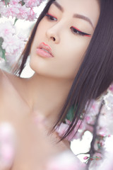 Outdoor fashion photo of beautiful young asian woman surrounded by flowers on spring. Perfect model with creative vivid makeup and pink lipstick on lips and traditional japanese hairstyle posing