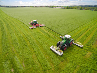 
Aerial view of a farmer in a modern tractor mowing a green fresh grass field on a sunny day with blue sky. 
