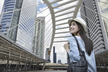 Young asian woman traveler with a backpack on her shoulder walking on pathway bridge over modern...