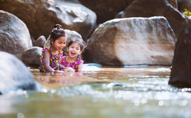 Two asian little child girls sitting on the rock and playing water in the riverside together in summer time with fun and happiness