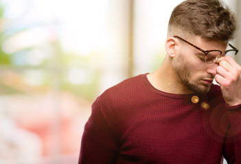 Handsome young man with sleepy expression, being overworked and tired, rubbes nose because of...