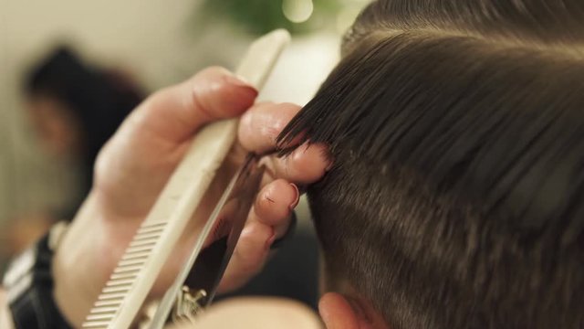 Haircutter combing hair and cutting with hairdressing scissors in barbershop. Close up hand hairdresser cutting wet male hair with hairdressing scissors in beauty salon.