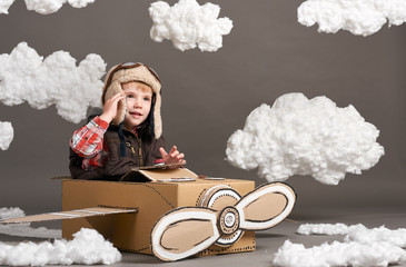 the boy plays in an airplane made of cardboard box and dreams of becoming a pilot, clouds of cotton wool on a gray background