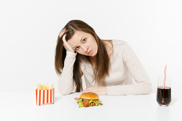 Sad tired woman put hand on head sitting at table with burger, french fries, cola in glass bottle isolated on white background. Proper nutrition or American classic fast food. Area with copy space.