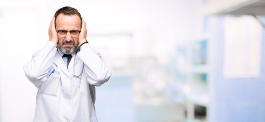 Obraz premium Doctor senior man, medical professional covering ears ignoring annoying loud noise, plugs ears to avoid hearing sound. Noisy music is a problem. at hospital