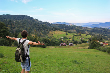 Guy with a backpack is standing on the hill. Beautiful mountains view with tall pines on background. Summer landscape with the village. Lifestyle travelling photo for young hipster.