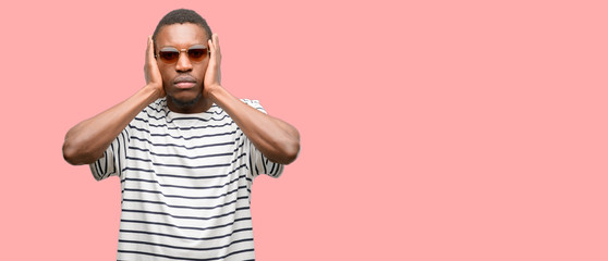 African black man wearing sunglasses covering ears ignoring annoying loud noise, plugs ears to avoid hearing sound. Noisy music is a problem.