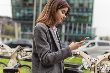 Happy girl with cell phone near skyscraper, young stylish woman using bicycle rent mobile app smiling outdoors, portrait of female manager browsing smartphone standing near bike sharing to go cycling