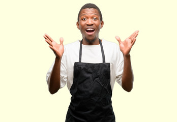 African man shop owner wearing apron happy and surprised cheering expressing wow gesture