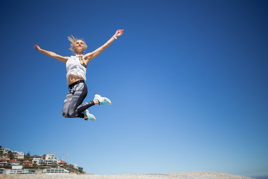 Beautiful blond female fitness model jumping for joy against a bright blue sky