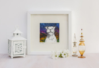 White interior display. Drawing of a happy Staffy dog with a big smile, on a collaged background in frame. With egyptian glass scent bottles, tea light and cherry blossom.