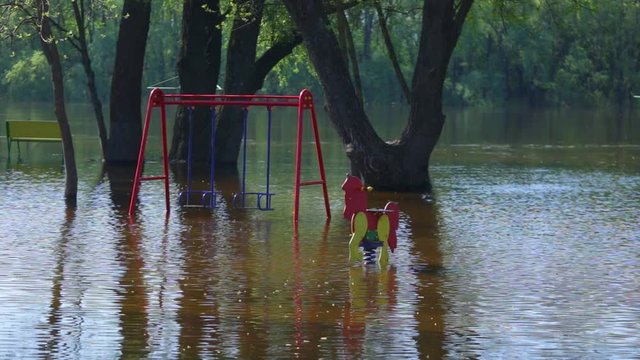 Flooded with river water empty spring beach with benches and green trees and children playground outdoors on sunny morning.