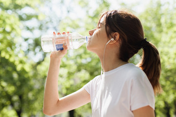 Brunette teenager girl drinking cold water after running in the park in the fresh air during sunny day, beautiful fitness athlete woman drinking water after work out, health and sport, flare light
