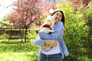 Naklejka premium Portrait of attractive young woman hugging cute jack russell terrier puppy in park, green lawn, foliage background. Hipster female in sunglasses smiling, pets her dog on grass. Close up, copy space.