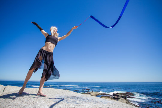 Beautiful female fitness model doing ribbon dancing on a granite rock overlooking the ocean on a bright and sunny summer day