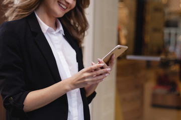 Smiling beautiful woman walking along the street using smartphone and doing online shopping, happy girl passing by a clothes shop window while browsing internet on her mobile phone and buying