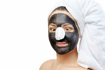  close-up portrait funny beautiful woman with facial black mask on white background, girl with a white towel on her head, satisfied and happy  smile, pampering, comic situation