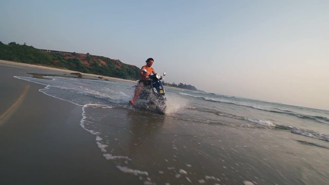 Scooter rides on a sandy beach in the sea splashing water.