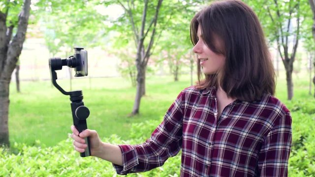 Vlogger with mobile phone camera stabilizer recording outdoor video blog. A  young pretty girl uses smartphone handheld gimbal for travel vlogging