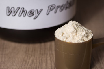 Whey Protein. Golden scoop with vanilla flavour powder and black jar of supplement. Wooden table. Close up, texture.