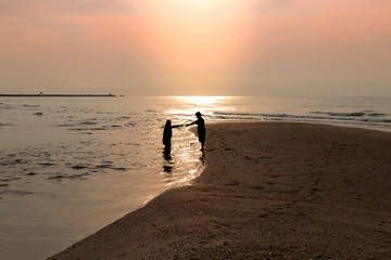 Silhouette of the couple held hands together at the seashore. Reflection of the sun on water surface. During in the morning with warm sunlight.