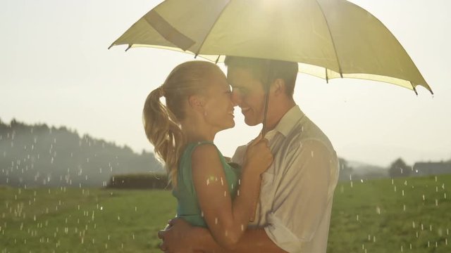 SLOW MOTION CLOSE UP LENS FLARE: Happy Caucasian couple on delightful date kisses under yellow umbrella. Ecstatic boyfriend and girlfriend enjoy the picturesque countryside on a rainy summer afternoon
