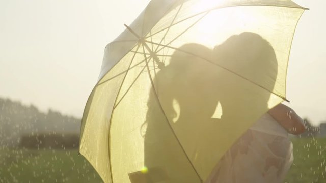 SLOW MOTION, CLOSE UP, LENS FLARE: Man and woman in love hide behind umbrella as they kiss in the golden countryside. Young Caucasian couple dancing in the rain stops and kisses during romantic date.