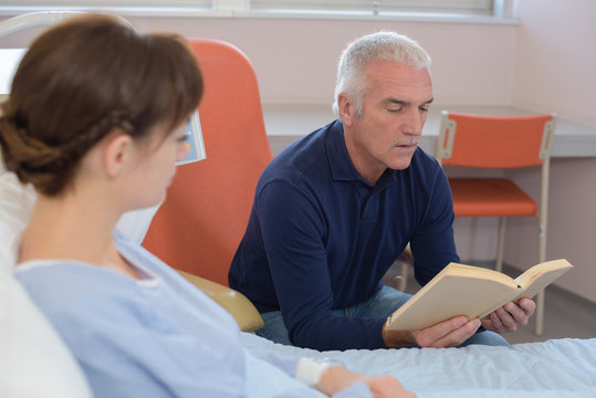 Man reading book to patient in hospital bed