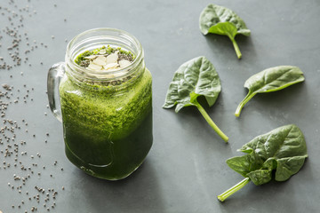 Healthy, green smoothie with spinach and almonds in a jar next to spinach leaves lying on a gray table