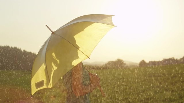 SLOW MOTION, CLOSE UP, SUN FLARE: Happy woman twirls in the spring rain while holding her yellow umbrella. Carefree girl with a blonde ponytail smiles and dances in sunny countryside in spite of rain.