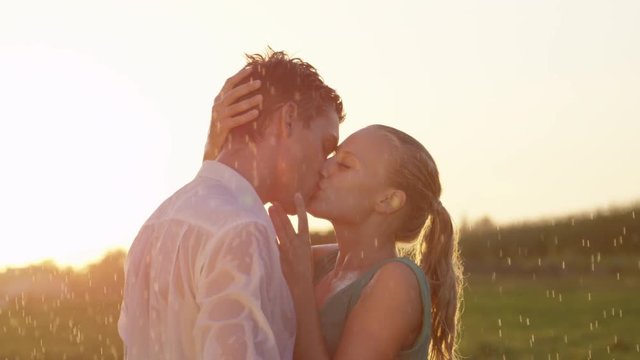 SLOW MOTION, CLOSE UP, LENS FLARE: Blonde woman gently caresses her boyfriend's face as they kiss in the rain. Young Caucasian couple gets wet during their romantic date in breathtaking summer nature.