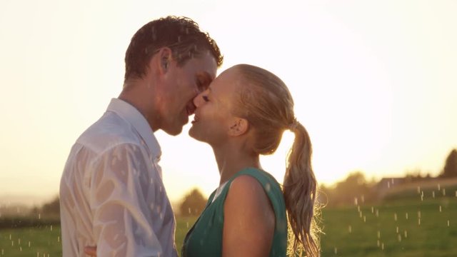 SLOW MOTION, CLOSE UP, LENS FLARE: Young Caucasian couple kisses in the rain on a beautiful spring morning. Smiling boyfriend and girlfriend kiss during delightful date in breathtaking summer nature.