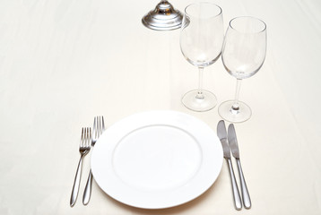 Dinner place setting in restaurant, copy space. Empty white plate with silver fork and knife, transparent wineglasses on beige tablecloth, free space for text. Elegant table setting in restaurant