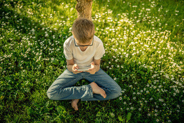 Young boy alone playing with smartphone. Pre teen with glasses sitting outdoor play with phone in hand. The use of the smartphone isolates the boy from the surrounding reality.