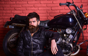 Hipster, brutal biker on pensive face in leather jacket sits, leans on motorcycle. Bikers lifestyle concept. Man with beard, biker in leather jacket near motor bike in garage, brick wall background.