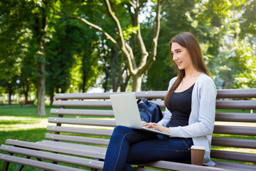 Obraz premium Motivated freelancer in the park. Girl on a bench, drinking coffee and working with laptop