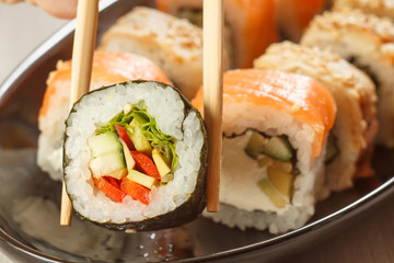Two chopsticks holding Hosomaki with vegetables and different  sushi rolls with seafood on ceramic plate on the background
