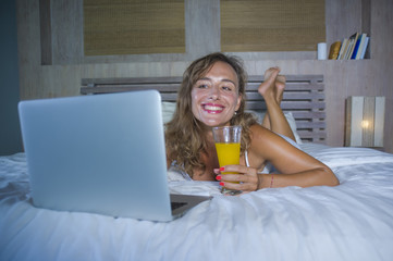 beautiful Caucasian woman 30s lying on bed at night in home bedroom using internet at laptop computer drinking healthy orange juice