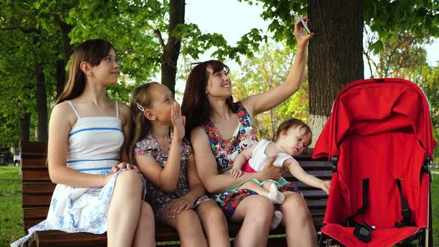 Young mother is photographed with her children on phone while sitting on bench in park and smiles at little baby sitting in stroller. Family makes selfie on phone and waving.