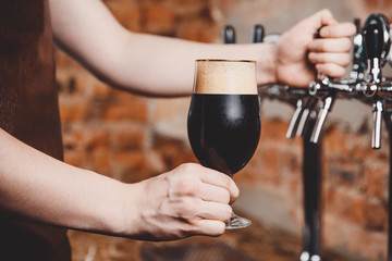 Barman holds in his hand dark unfiltered cold beer is poured into glass beaker against background of taps for bottling.